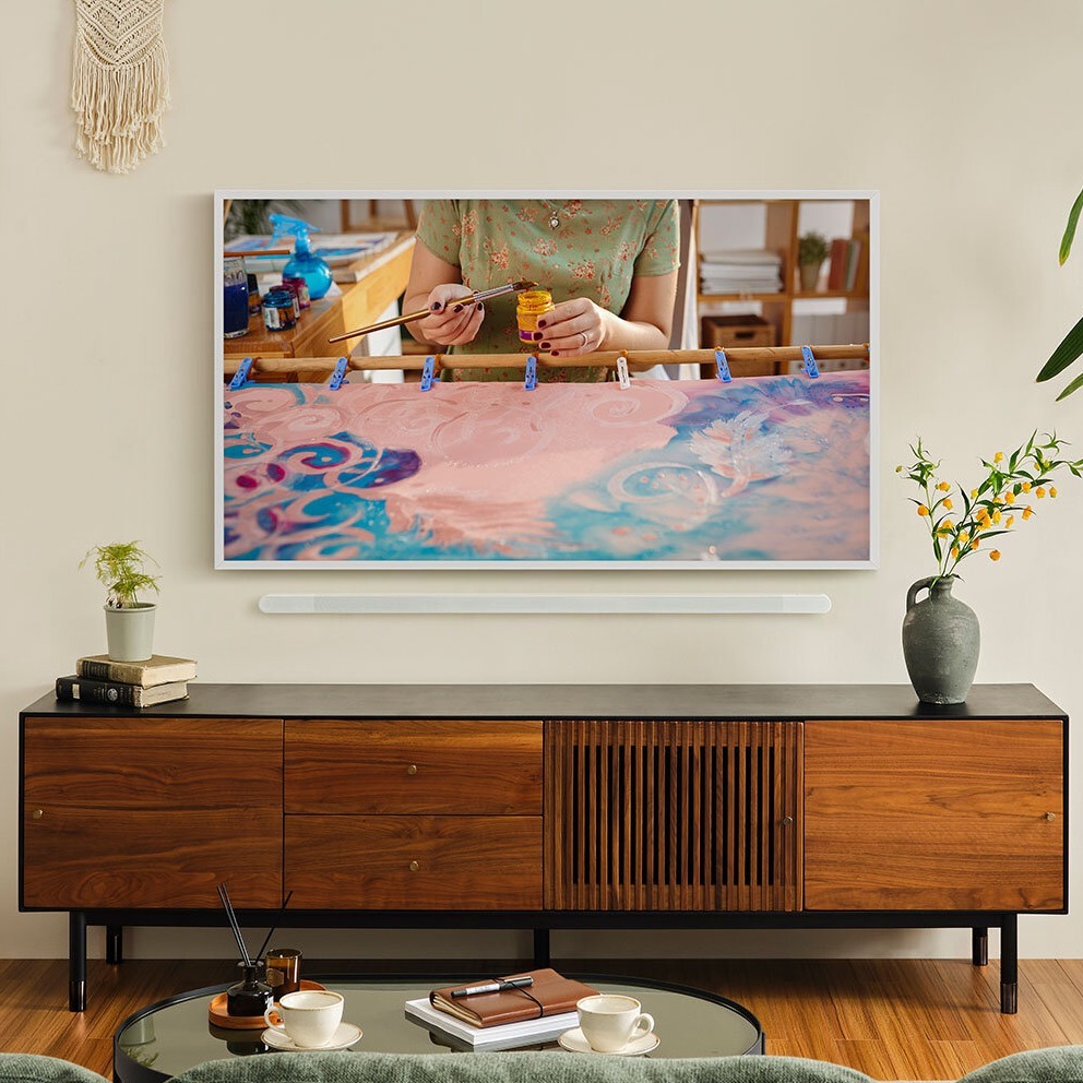 Woman on sofa in front of LG brand QNED smart TV, using TV remote to select entertainment option from the TV's home screen