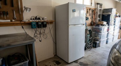 Top mount fridge in a garage, plugged into a power board that also has other items plugged into it. Image generated by AI