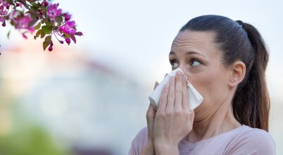 A woman with hayfever allergies is covering her nose with a tissue and looking at a flowering tree