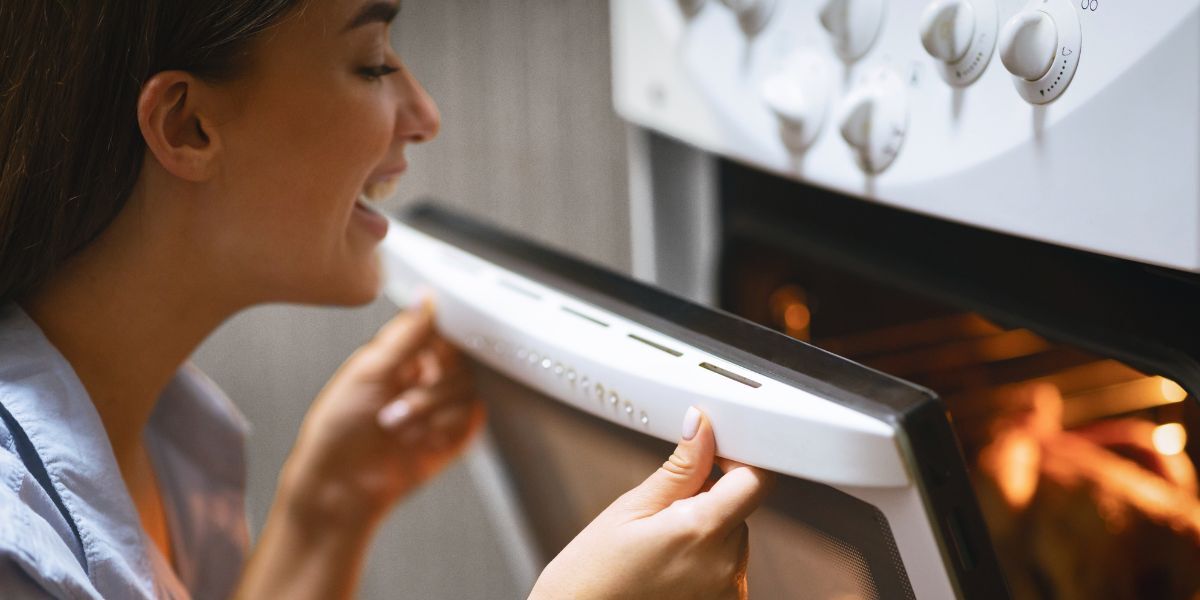 A smiling woman opens an oven to look at the dish cooking inside